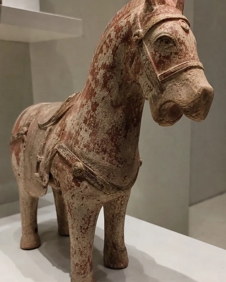 A weathered terracotta horse statue displayed on a white platform in a museum setting.