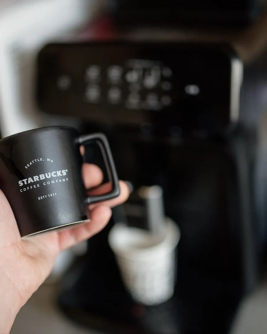 A hand holding a black ceramic mug in front of a blurry espresso machine.