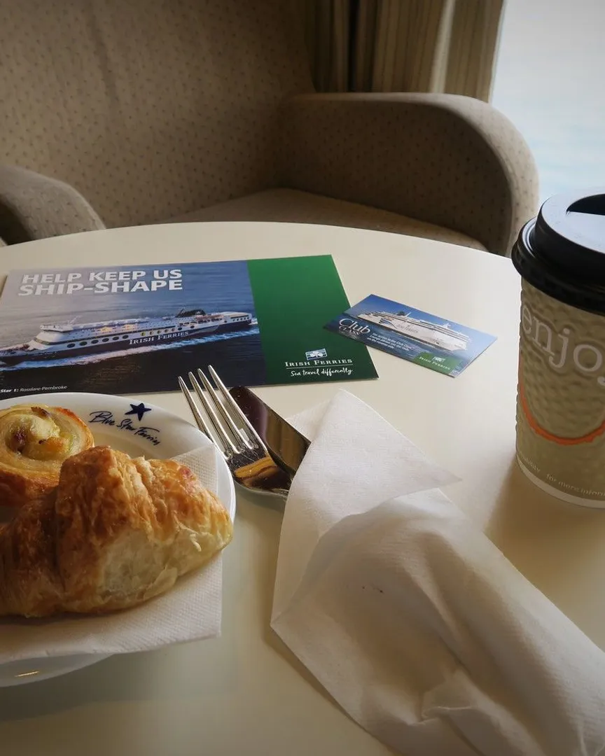 A croissant and coffee cup on a table inside a ferry lounge with travel brochures.