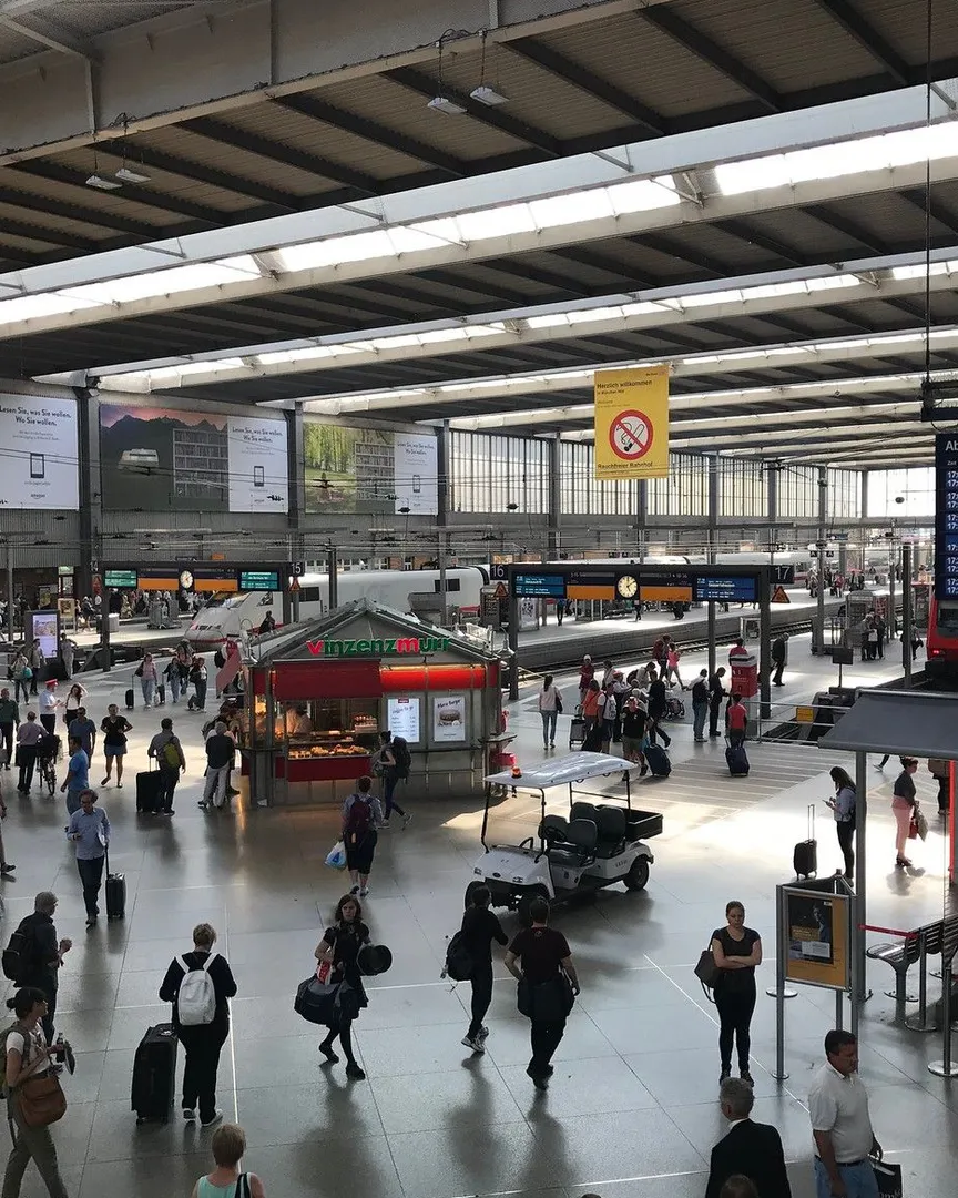 A busy train station terminal with travelers, a food kiosk, and trains in the background.