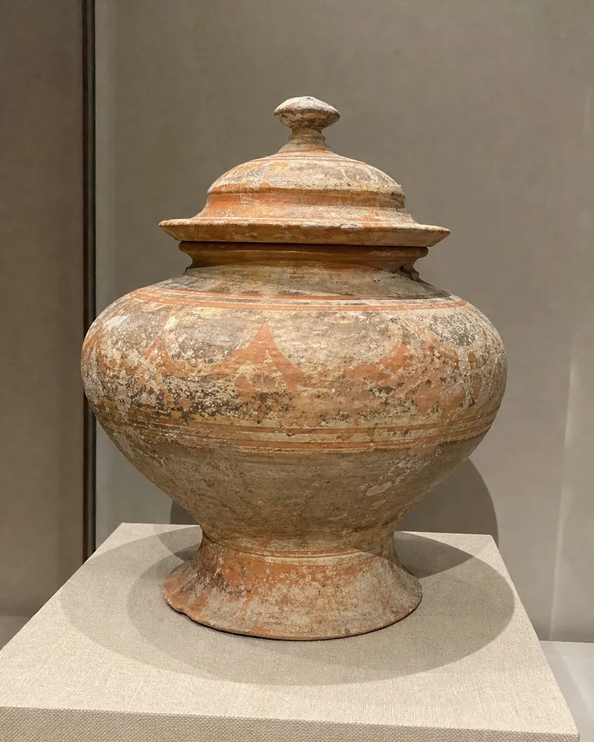 An ancient painted ceramic jar with a lid on a museum display pedestal.