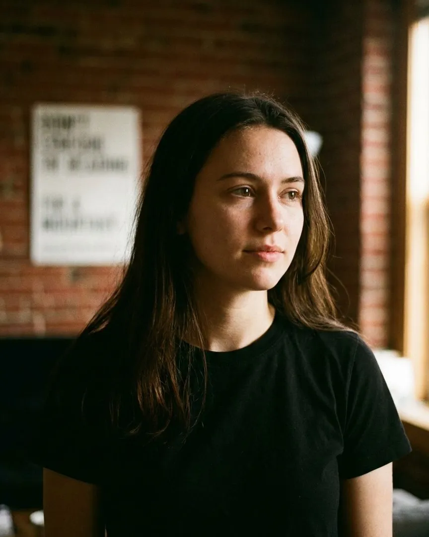 A young woman with long dark hair wearing a black t-shirt in front of a brick wall.