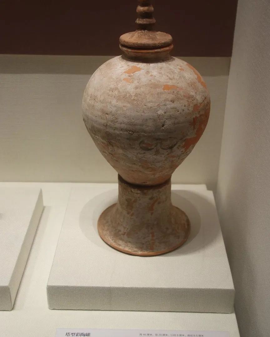 An ancient painted pottery jar with a lid displayed on a pedestal in a museum.