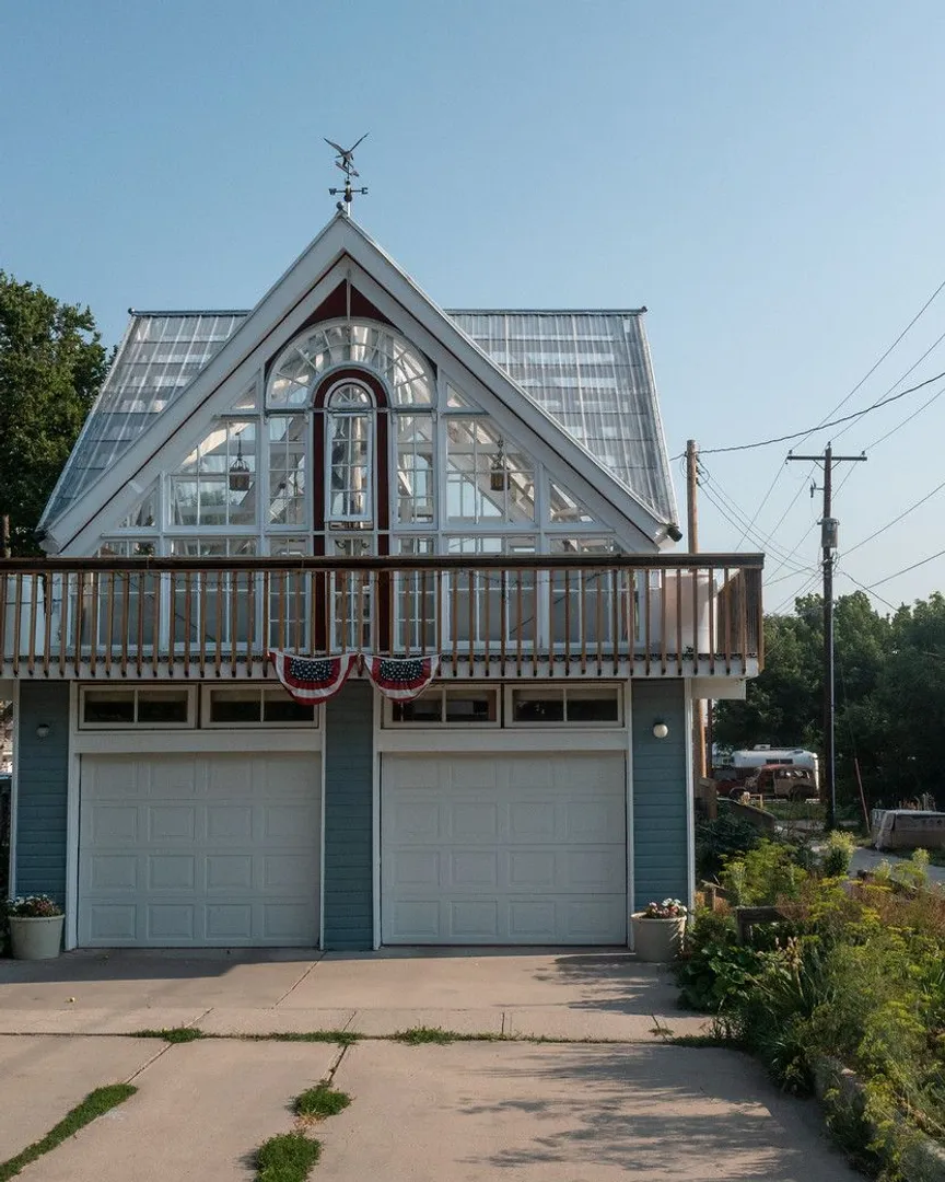 A two-story blue garage with a glass greenhouse on the upper level and a wooden balcony.