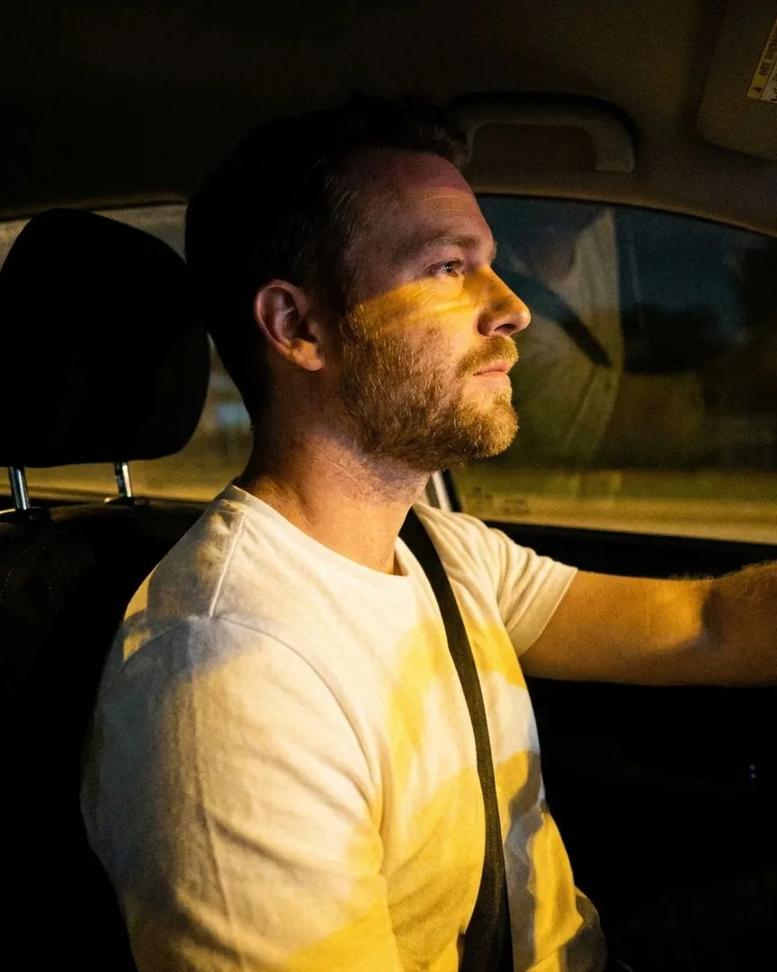 A man with a beard driving a car at night with yellow light on his face.
